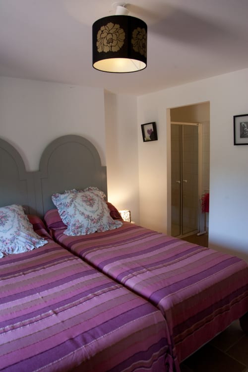 Chambre aux Chambres d'hôtes Les Pratges à Figeac, avec literie à rayures violettes, oreillers à motifs floraux et luminaire noir en forme de fleur au plafond