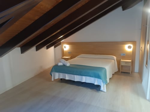 Moderno dormitorio abuhardillado en Apartamentos Turísticos Ávila con cama queen, vigas de madera, iluminación lateral y suelo de madera