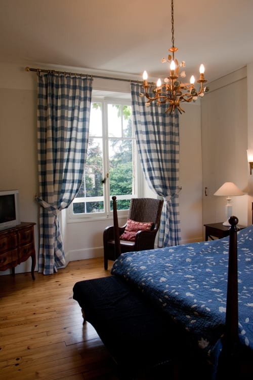 Chambre élégante aux Chambres d'hôtes Les Pratges avec rideaux à carreaux bleus, lustre en laiton et lit en bois