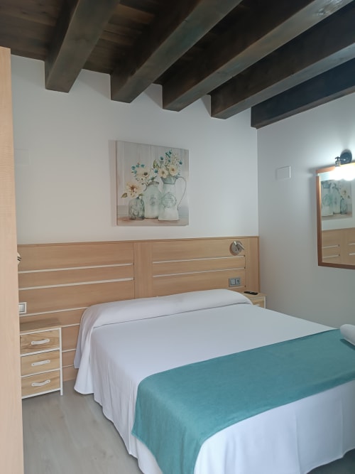 Dormitorio moderno en Apartamentos Turísticos Ávila con cama de madera, ropa de cama blanca, manta turquesa, vigas vistas y detalles florales