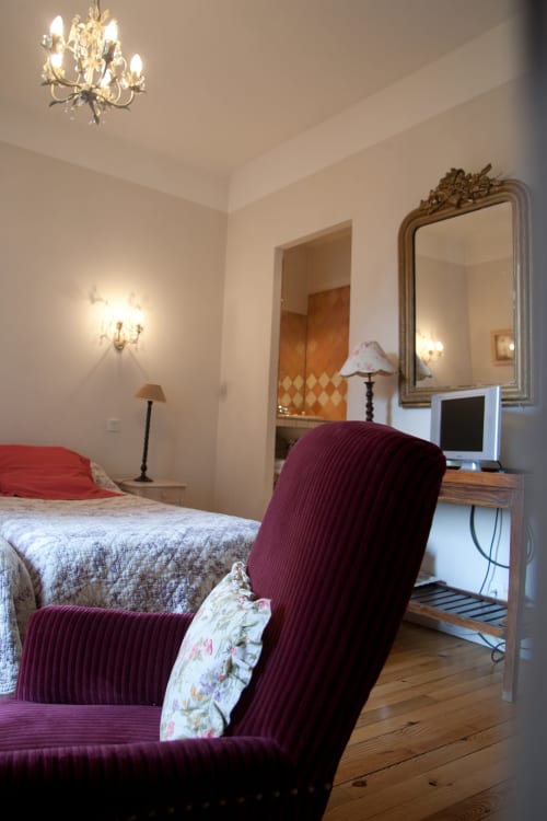 Chambre élégante aux Chambres d'hôtes Les Pratges à Figeac : fauteuil bordeaux, lit, miroir travaillé et lustre en cristal