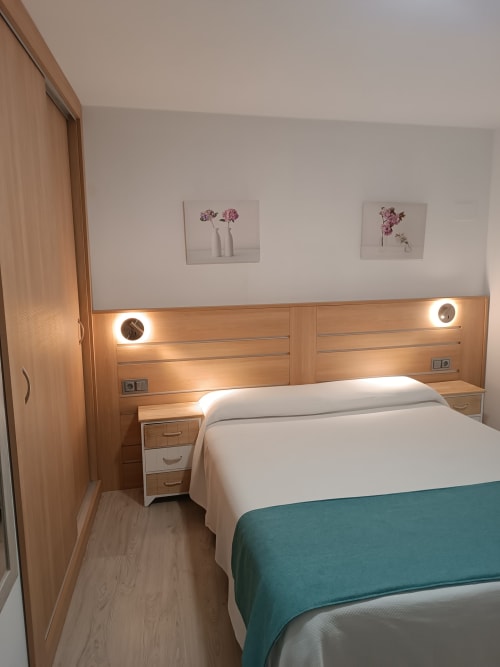 Dormitorio moderno en Apartamentos Turísticos Ávila con cabecero de madera, ropa de cama blanca, manta turquesa y cuadro con flores rosadas