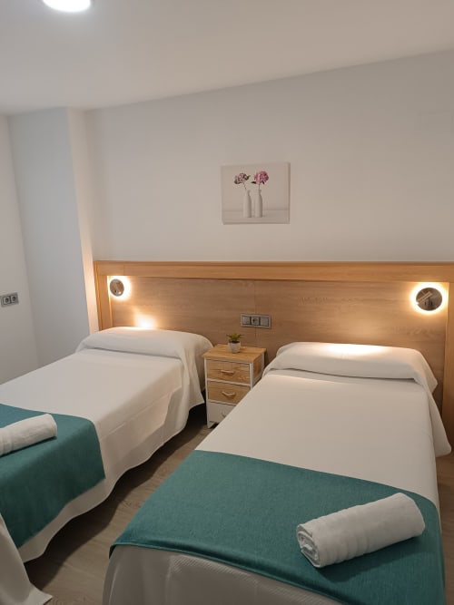 Habitación doble en Apartamentos Turísticos Ávila con sábanas blancas, mantas azul turquesa, cabecero de madera y cuadros de flores rosas