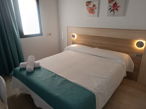 Moderno dormitorio en Apartamentos Turísticos Ávila con cama blanca, cabecero de madera, luces LED y ventana con vista a la ciudad