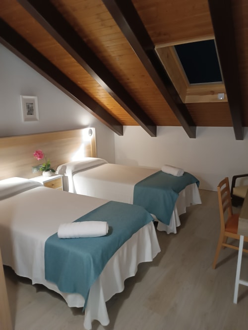 Dormitorio doble en Apartamentos Turísticos Ávila con vigas de madera, paredes blancas, mantas azul verdoso y lucernario