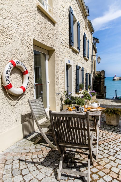 Terrasse côtière de la maison d'hôtes Les Filles du Bord de Mer à Port-en-Bessin avec mobilier de jardin, fleurs et vue sur l'océan.