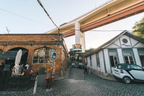 Pátio do VAGO Alcântara Suites com café de tijolos, carro elétrico branco e moderno viaduto em Lisboa