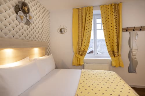 Chambre standard à l'Hôtel De Lion Sur Mer avec tête de lit matelassée blanche, rideaux à motifs jaunes et équipements modernes.