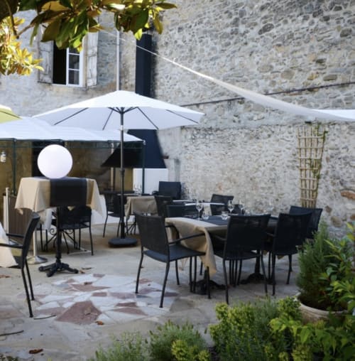 Patio de piedra con área de comedor en Domaine de Bize: sombrilla, mesas y sillas negras en Mirepoix