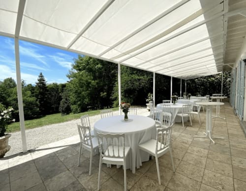Terrasse d'événement en plein air avec store rétractable blanc, tables rondes et chaises donnant sur les jardins du Domaine de Bize