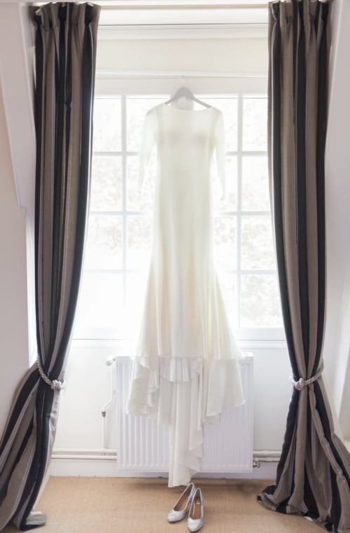 Elegantes weißes Hochzeitskleid hängend am Fenster mit grauen Vorhängen im Domaine de Bize Mirepoix