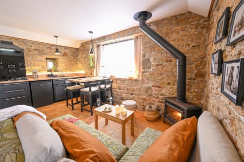 Cozy open-plan living space at Espace Ker Gana B&B in Plourin featuring stone walls, wood stove, sofa, and modern kitchen.