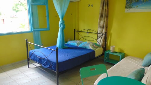 Chambre lumineuse aux murs jaune vif chez Caribgites à Sainte-Anne, avec lit métallique bleu, vue sur le jardin et accents turquoise