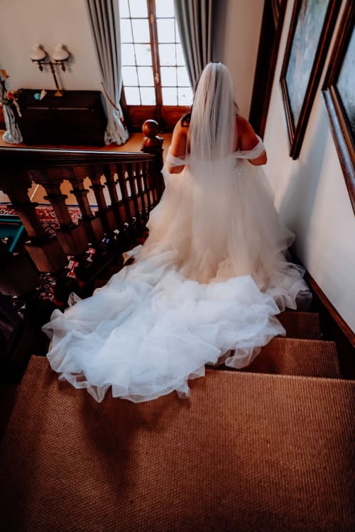 Braut im weißen Kleid mit langem Schleier auf der Holztreppe des Domaine de Bize, Hochzeitsvenue in Mirepoix