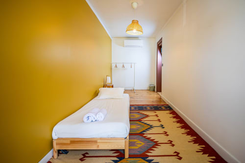 Quarto moderno no VAGO Alcântara Suites em Lisboa com parede de destaque amarela, cama de solteiro, piso de madeira e tapete colorido