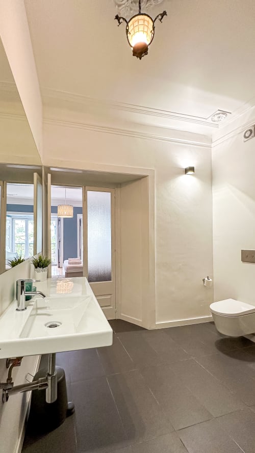 Casa de banho moderna com lavatório branco, duche, arandela de parede e piso em mosaico escuro no VAGO Alcântara Suites Lisboa
