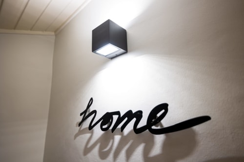 Luminária de parede cúbica preta moderna iluminando parede branca com texto 'home' no quarto do hotel VAGO Alcântara Suites, Lisboa