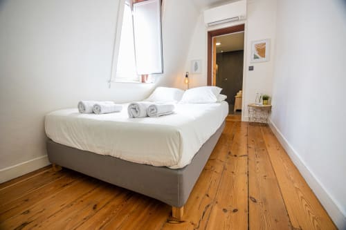 Quarto moderno nas Suites VAGO Alcântara em Lisboa com piso de madeira, cama cinzenta, almofadas brancas e toalhas enroladas