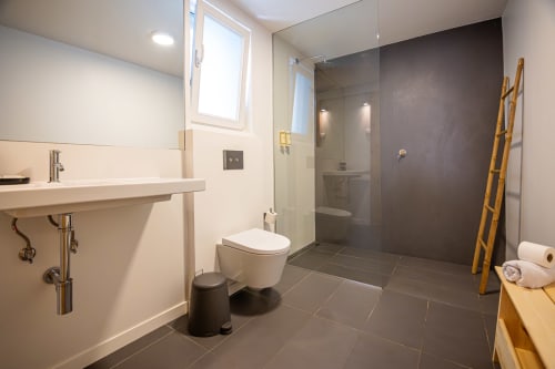 Banheiro moderno em VAGO Alcântara Suites Lisboa com chuveiro walk-in, vaso sanitário suspenso, pia branca e prateleira de escada em madeira