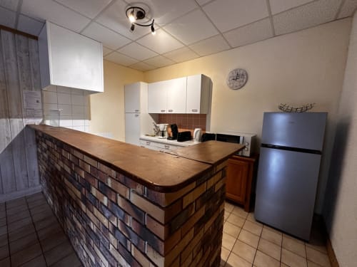 Îlot de cuisine en brique avec plan de travail en bois, armoires blanches, réfrigérateur et ventilateur au plafond aux Gîtes Des Planchettes