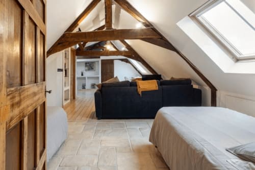 Chambre mansardée rustique à Villa République à Caen avec poutres apparentes, lucarne, canapé noir et carrelage en pierre.
