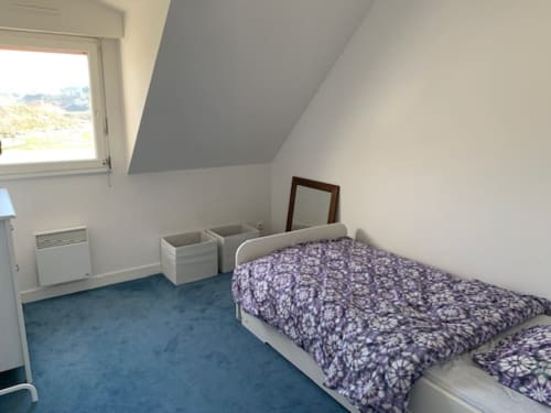 Chambre mansardée lumineuse avec literie à motifs violets, moquette bleue, vue sur Belle-Île-en-Mer, radiateur et rangements