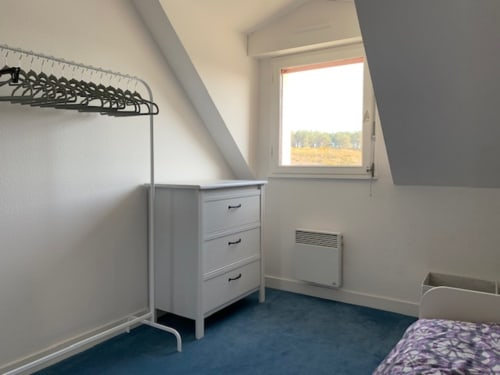 Chambre mansardée lumineuse à l'appartement Locmaria avec murs blancs, sol bleu, commode, portant et fenêtre