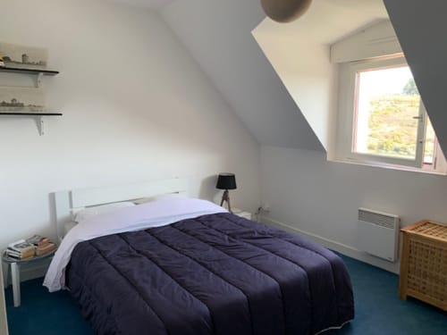 Chambre lumineuse sous les combles avec édredon bleu foncé, cadre de lit blanc, vue sur la mer et mobilier en bois à Loc'Maria Belle