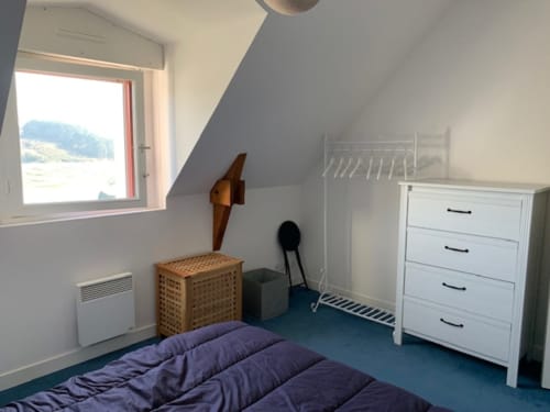 Chambre mansardée lumineuse avec vue sur mer, literie violette, commode blanche, panier de rangement en bois, mobilier minimaliste