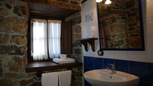 Baño rústico del Hotel Rural Los Texos con paredes de piedra, lavamanos blanco, detalles en azulejos azules y ventana con vistas al jardín