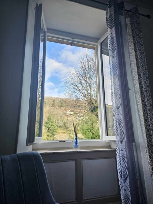 Offenes Fenster der Bastide du Thoré mit Garten- und Bergblick, gemusterte Gardinen, blaue Vase auf der Fensterbank