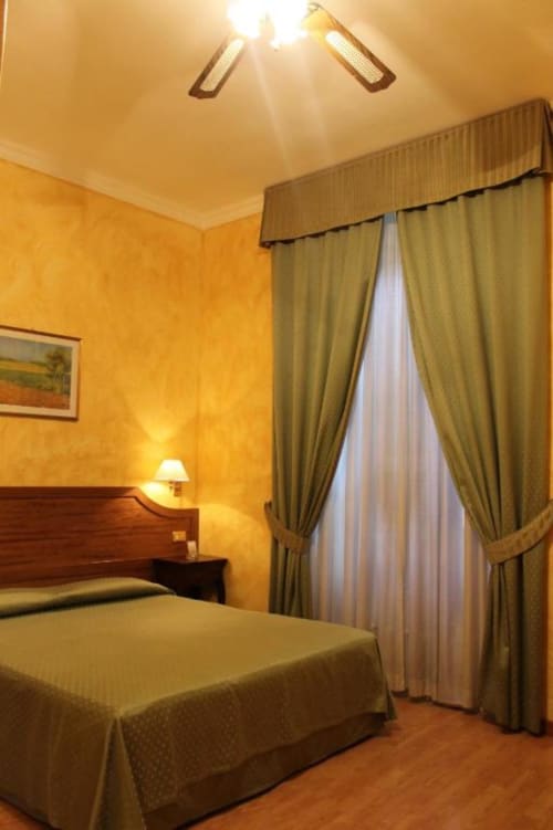 Elegante camera d'hotel calda con letti matrimoniali, tende color oliva, comodino in legno con lampada e parquet all'Hotel Fiori Roma