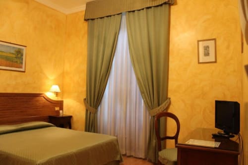 Accogliente camera d'hotel in tonalità gialle presso Hotel Fiori Roma con letto matrimoniale, tende verdi, scrivania, televisore e vista sulla città dalla finestra
