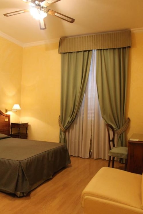 Ampia camera d'albergo presso Hotel Fiori Roma con letti gemelli, pareti gialle, tende color oliva, ventilatore a soffitto e pavimenti in legno