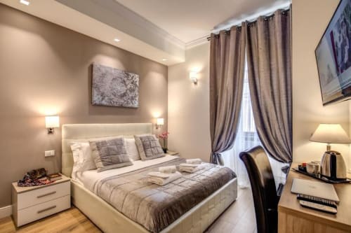 Camera da letto di Chester Suites Roma con letto in tessuto color crema, pareti beige, tende tortora e illuminazione moderna