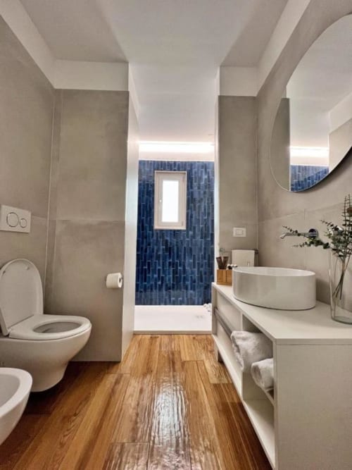 Bagno moderno a Finella Charming Rooms a Bari con sanitari bianchi, parete di piastrelle blu, pavimenti in legno e specchio rotondo