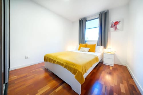 Dormitorio luminoso en Villa Cacemar con ropa de cama amarilla, suelos de madera, cortinas grises, mesilla blanca y vistas al mar