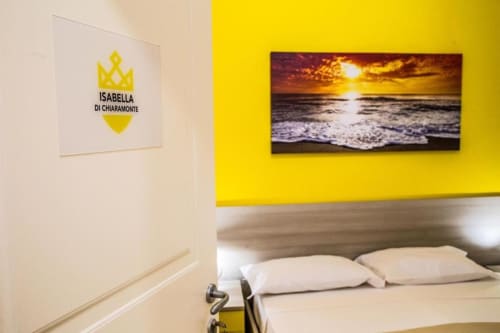 Dormitorio amarillo con logo Isabella di Chiaramonte, obra de arte playa atardecer y almohadas blancas en Borgo Angioino Naples B&B