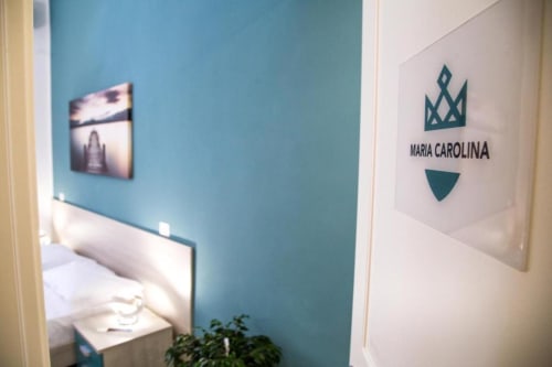 Ingresso della camera con pareti blu teal, segnaletica del logo Maria Carolina e opera d'arte con vista sulla città a Borgo Angioino Napoli