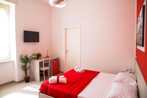Camera moderna presso Borgo Angioino BnB Napoli con muri rosa, letto rosso, scrivania, pianta e televisore schermo piatto