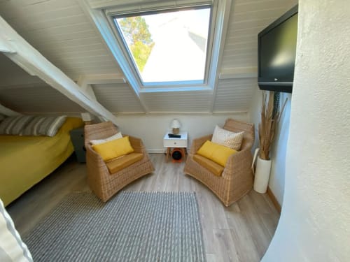 Chambre confortable mansardée avec deux fauteuils en osier, coussins jaunes, fenêtre de toit, TV et tapis rayé