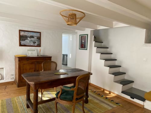 Salle à manger lumineuse avec bureau en bois, escalier et lampe en rotin aux Locations Loc'Maria, Belle Île en Mer