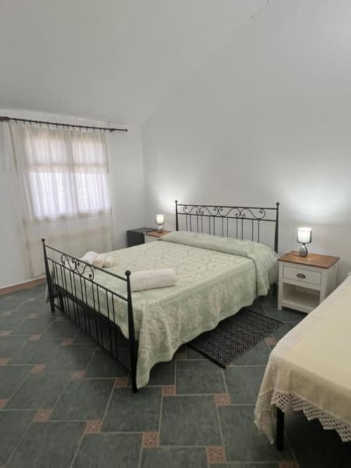 Camera luminosa all'Agriturismo Su Barraccu con letto in ferro battuto, biancheria bianca, comodini e finestre con vista sul giardino