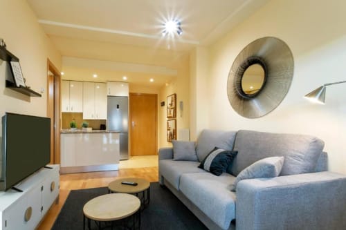 Moderno salón de apartamento con sofá gris, espejo redondo, cocina y suelos de madera en Burgos