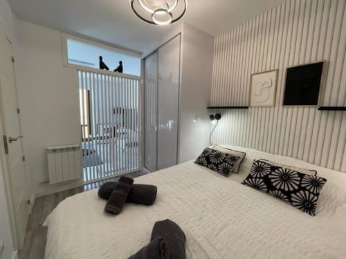 Moderno dormitorio en el apartamento Urban Home de Burgos con ropa de cama blanca, cojines decorativos negros, TV montado en pared y
