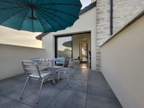 Patio moderne des Gîtes Les Camelias 71 à Oslon avec parasol turquoise, chaises métalliques et portes vitrées ouvertes sur l'intérieur