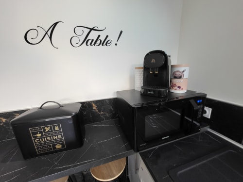 Petite kitchenette moderne aux Gîtes Les Camelias : plan de travail en marbre noir, cafetière, micro-ondes et sticker mural « A Table !»