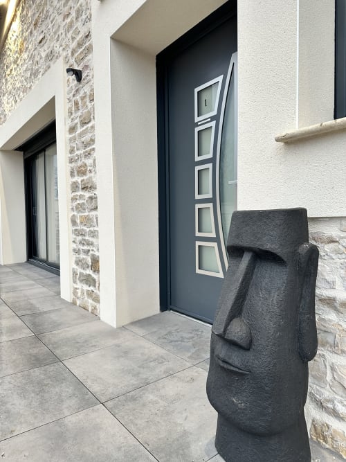 Entrée contemporaine des Gîtes Les Camelias 71 avec porte sombre, détails en pierre et sculpture moai moderne dans le jardin d'Oslon