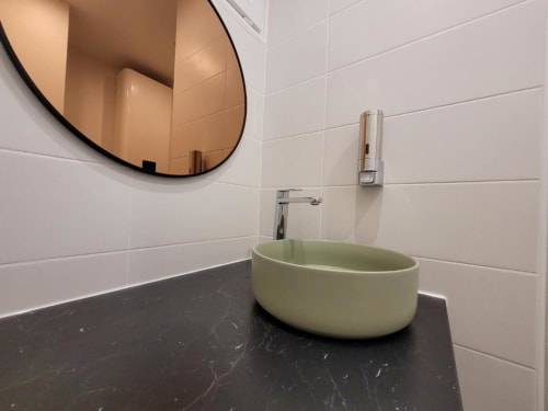 Lavabo moderne aux teintes vert pâle sur plan de travail en marbre noir, miroir rond, carrelage blanc à l'Appart Hôtel