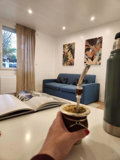 Main tenant un yerba maté dans l'appartement Appart Hôtel Panambi avec canapé bleu, livre ouvert et vue sur la ville